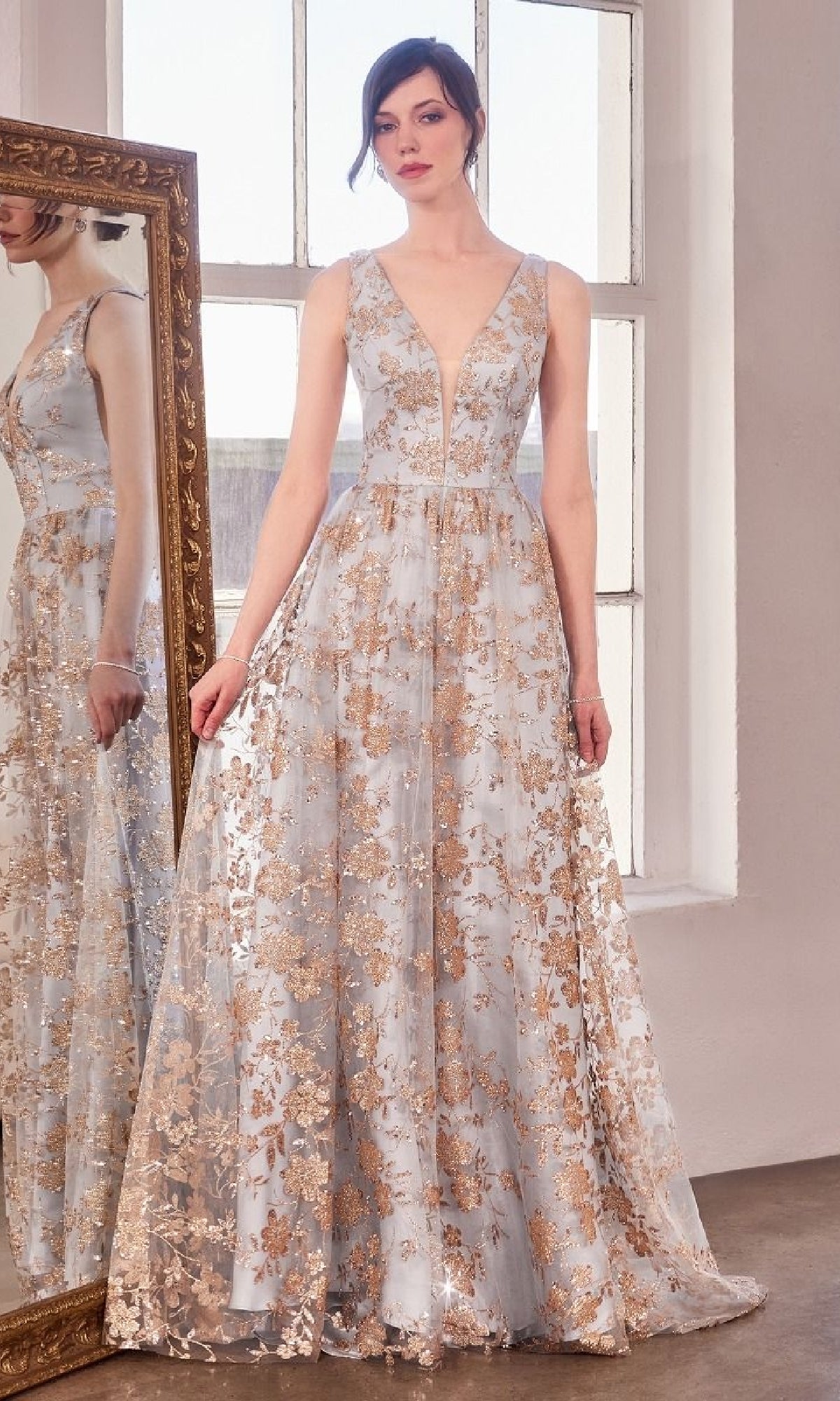 A woman stands indoors in front of a tall mirror, wearing the Amoris T606 Embroidered-Lace Long Prom Dress with a deep V-neck and gold floral details, gazing slightly to the side.